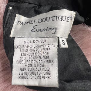 K&ouml;nnte beinhalten: Ein wei&szlig;es Etikett mit schwarzem Text, auf dem "PAPELL BOUTIQUE Evening" steht und die Stoffzusammensetzung des Kleidungsst&uuml;cks beschrieben wird. Das Etikett ist an einem schwarzen Kleidungsst&uuml;ck mit einem Rei&szlig;verschluss befestigt. Das Etikett lautet "SHELL: 100% SILK EXCLUSIVE OF ORNAMENTATION LINING 100% POLYESTER EXTERIEUR 100% SOIE SAUF LES ORNEMENTS DOUBLURE 100% POLYESTER MADE IN INDIA FABRIQUER AUX INDES SEE REVERSE FOR CARE INSTRUCTIONS AU VERSO" und hat ein Gr&ouml;&szlig;enetikett "S".