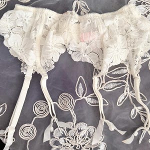 Puede incluir: Liga de encaje marfil con detalles florales. La liga presenta tirantes ajustables y un delicado borde festoneado. El fondo es una tela transparente con patrones florales bordados.