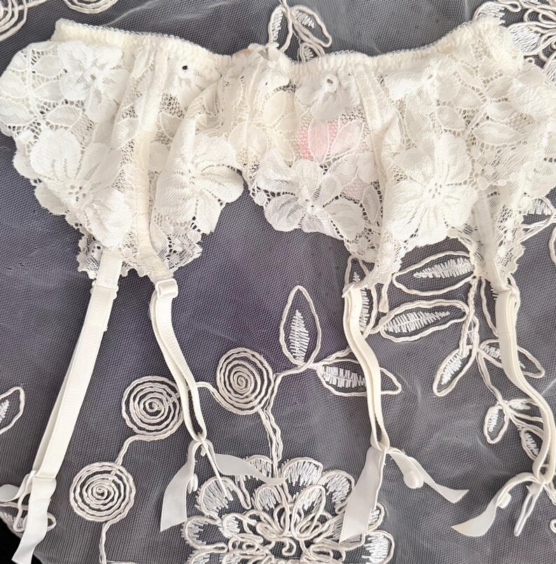 Puede incluir: Liga de encaje marfil con detalles florales. La liga presenta tirantes ajustables y un delicado borde festoneado. El fondo es una tela transparente con patrones florales bordados.