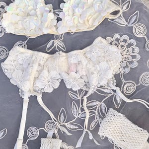 Puede incluir: Un conjunto de lencer&iacute;a blanco con un sujetador con lentejuelas y una liga con detalles de encaje floral. El conjunto incluye una liga a juego y un peque&ntilde;o accesorio en forma de red. Los art&iacute;culos est&aacute;n dispuestos sobre una tela blanca bordada con motivos florales.