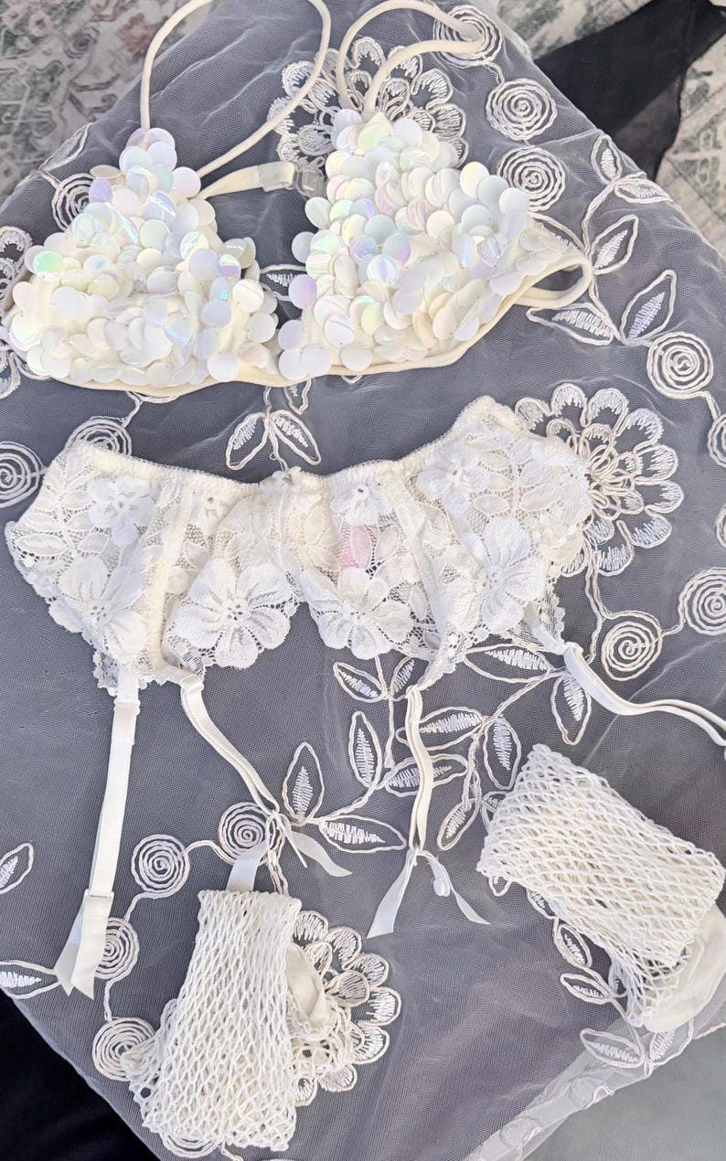 Puede incluir: Un conjunto de lencer&iacute;a blanco con un sujetador con lentejuelas y una liga con detalles de encaje floral. El conjunto incluye una liga a juego y un peque&ntilde;o accesorio en forma de red. Los art&iacute;culos est&aacute;n dispuestos sobre una tela blanca bordada con motivos florales.