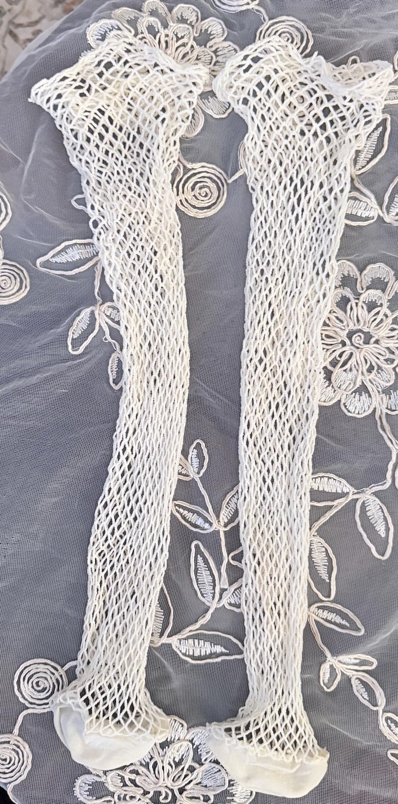 Puede incluir: Un par de medias de color crema con tejido abierto. Las medias est&aacute;n hechas de un material similar a una red y se muestran sobre un fondo de tela bordada blanca con motivos florales y de hojas. Las medias son un accesorio de moda.