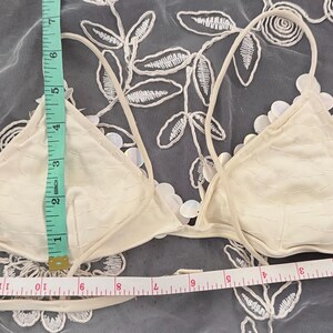 Puede incluir: Top de bikini triangular de color marfil con detalles florales. El top mide aproximadamente 15 cm de alto y 30 cm de ancho. Tiene tirantes finos y ajustables. El top de bikini se muestra sobre un fondo blanco con estampado floral.