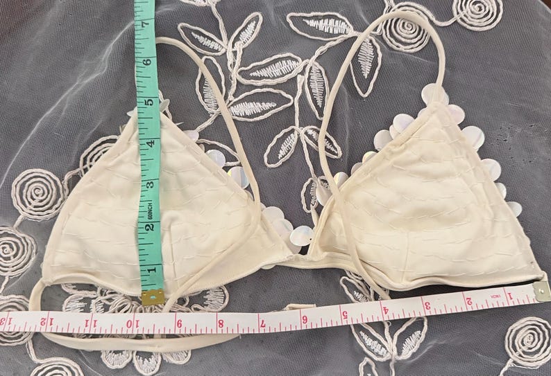 Puede incluir: Top de bikini triangular de color marfil con detalles florales. El top mide aproximadamente 15 cm de alto y 30 cm de ancho. Tiene tirantes finos y ajustables. El top de bikini se muestra sobre un fondo blanco con estampado floral.