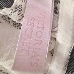 Puede incluir: Primer plano de una etiqueta rosa con las palabras "VICTORIA'S SECRET" impresas en un rosa m&aacute;s oscuro. La etiqueta est&aacute; adherida a una tela de encaje blanco. El fondo es una malla gris oscuro.