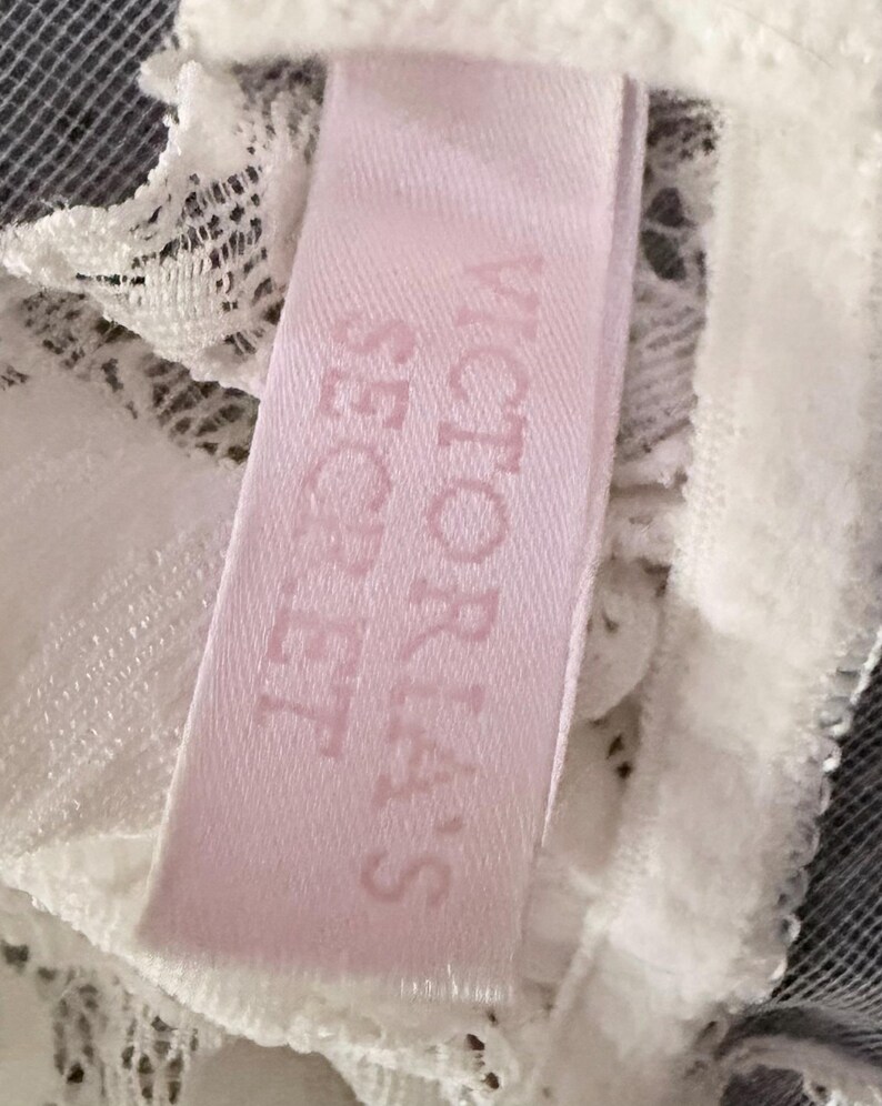 Puede incluir: Primer plano de una etiqueta rosa con las palabras "VICTORIA'S SECRET" impresas en un rosa m&aacute;s oscuro. La etiqueta est&aacute; adherida a una tela de encaje blanco. El fondo es una malla gris oscuro.