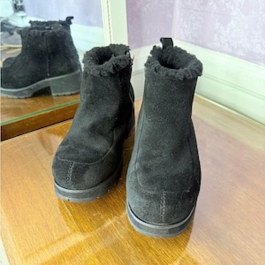Könnte beinhalten: Ein Paar schwarze Wildleder-Ankle Boots mit Plateausohle. Die Stiefel haben ein Futter aus Kunstfell und einen Reißverschluss. Die Stiefel spiegeln sich in einem Spiegel. Die Stiefel sind etwa 15 cm hoch.