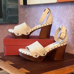 May include: A pair of cream-colored platform sandals with a textured upper and wooden heels. The sandals feature an ankle strap with a buckle and are adorned with small, round studs. The heels are approximately 4 inches high.