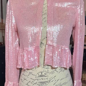 May include: A light pink sequined jacket with long sleeves and a high collar. The jacket features a ruffled hem and cuffs. The garment is displayed on a mannequin. The sequins create a shimmering effect.