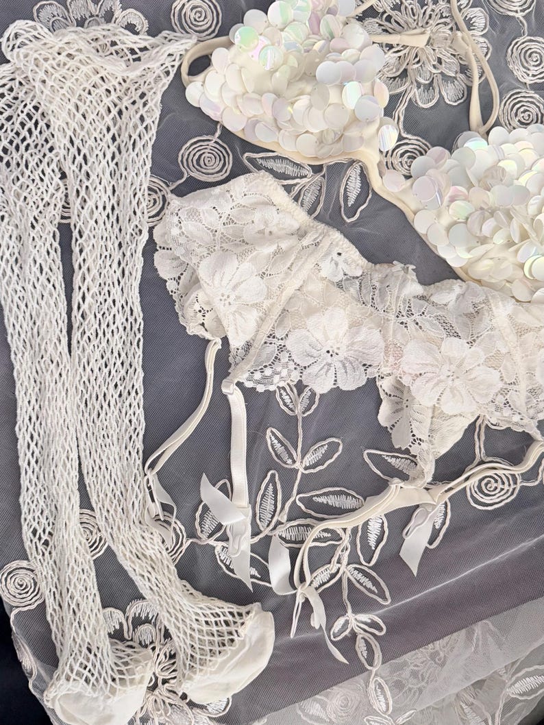 Puede incluir: Un surtido de lencer&iacute;a blanca, que incluye un sujetador adornado con lentejuelas iridiscentes, un sujetador de encaje y un par de medias de red. Los art&iacute;culos est&aacute;n dispuestos sobre una superficie gris con bordados florales.