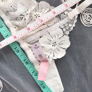 Puede incluir: Primer plano de un sujetador de encaje blanco con motivos florales. Se adjunta una etiqueta rosa y una cinta m&eacute;trica muestra las dimensiones del sujetador. El sujetador est&aacute; sobre una superficie gris.