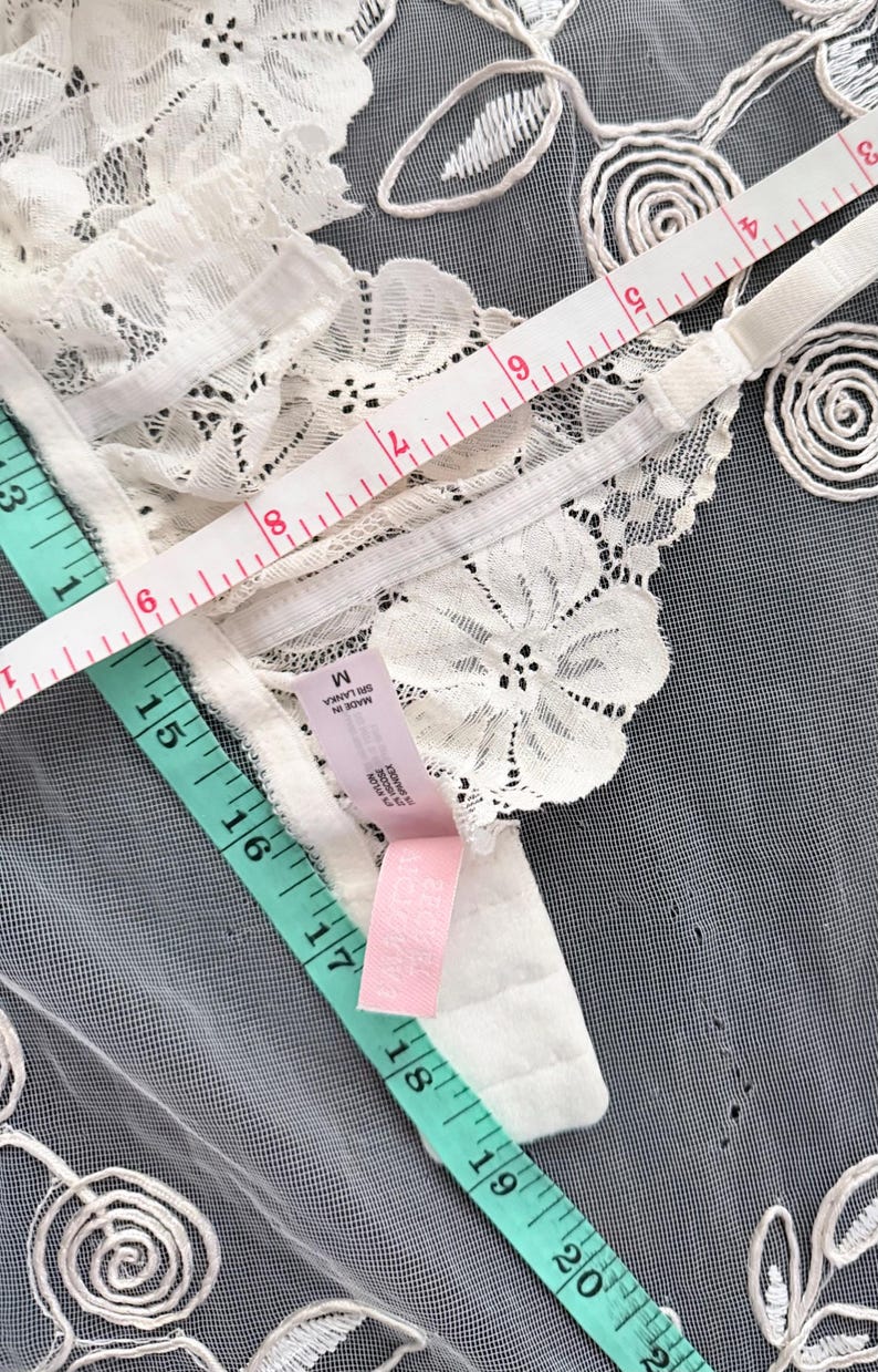 Puede incluir: Primer plano de un sujetador de encaje blanco con motivos florales. Se adjunta una etiqueta rosa y una cinta m&eacute;trica muestra las dimensiones del sujetador. El sujetador est&aacute; sobre una superficie gris.