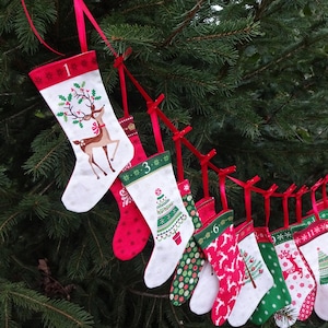 Puede incluir: Una cadena de 12 calcetines de Navidad con cinta roja y diferentes diseños navideños, incluyendo renos, árboles de Navidad y copos de nieve. Los calcetines están numerados del 1 al 12.