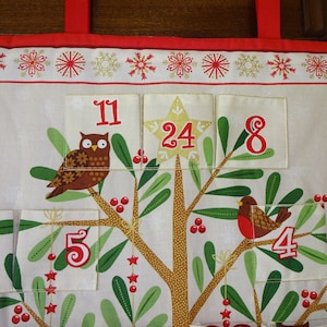 Puede incluir: Un calendario de adviento de tela blanca con un dise&ntilde;o de &aacute;rbol de Navidad. El &aacute;rbol tiene hojas verdes, un tronco marr&oacute;n y bayas rojas. Cada bolsillo tiene un n&uacute;mero, incluyendo 11, 24, 8, 23, 5, 4 y 3. Hay un b&uacute;ho en el &aacute;rbol y un petirrojo en una rama.