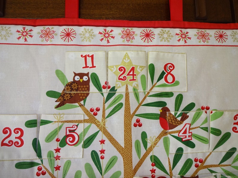 Puede incluir: Un calendario de adviento de tela blanca con un dise&ntilde;o de &aacute;rbol de Navidad. El &aacute;rbol tiene hojas verdes, un tronco marr&oacute;n y bayas rojas. Cada bolsillo tiene un n&uacute;mero, incluyendo 11, 24, 8, 23, 5, 4 y 3. Hay un b&uacute;ho en el &aacute;rbol y un petirrojo en una rama.