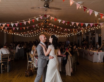 May include: A bride and groom dance at their wedding reception. The couple is surrounded by guests seated at tables decorated with white tablecloths and pink and white bunting. String lights hang above the dance floor.