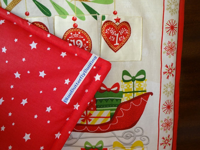 Puede incluir: Tela roja con estrellas blancas y una etiqueta azul que dice "BlueButterflyBunting". La tela est&aacute; doblada sobre una tela blanca con un dise&ntilde;o de calendario de adviento navide&ntilde;o. El calendario tiene un coraz&oacute;n rojo con el n&uacute;mero "1" en &eacute;l, un adorno rojo con el n&uacute;mero "19" en &eacute;l y un trineo rojo con regalos.