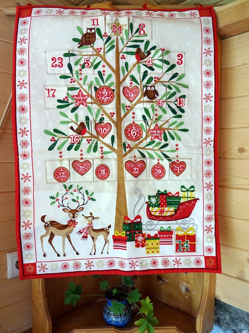 Puede incluir: Un calendario de adviento de tela blanca con forma de &aacute;rbol de Navidad con detalles rojos, verdes y dorados. El calendario presenta los n&uacute;meros del 1 al 24, b&uacute;hos, renos y un trineo con regalos.