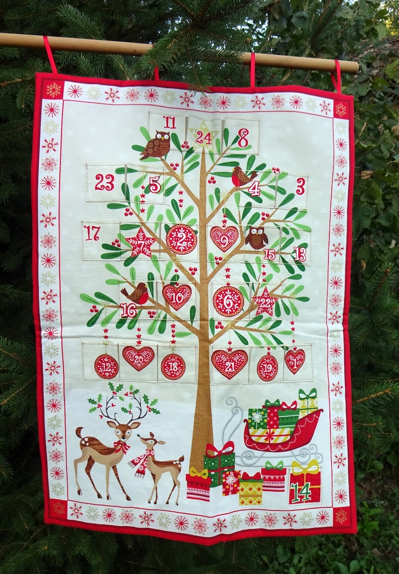 Puede incluir: Un calendario de adviento de tela blanca con forma de &aacute;rbol de Navidad con detalles rojos, verdes y dorados. El &aacute;rbol tiene 24 bolsillos numerados, cada uno con un dise&ntilde;o festivo diferente. Hay dos ciervos en la parte inferior del calendario y un trineo con regalos.