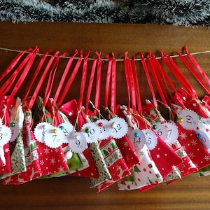 Puede incluir: Un calendario de adviento navideño hecho a mano con 24 pequeñas bolsas de tela, cada una numerada y colgada de una cuerda. Las bolsas son rojas con diseños festivos y atadas con cintas rojas. El calendario está decorado con guirnaldas verdes y plateadas.