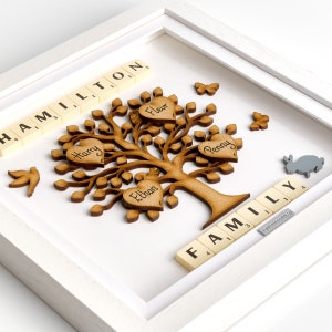 May include: A white frame with a wooden family tree inside. The tree has brown leaves and branches. There are five hearts on the tree, each with a name written on it: Fleur, Harry, Ethan, Penny. The words "HAMILTON" and "FAMILY" are spelled out with white tiles below the tree.