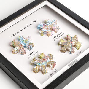 May include: A framed display featuring four 3D puzzle pieces printed with detailed map sections. The text reads Dominic & Danielle, Met 10th October 1992, Engaged 31st December 2002, Married, and Live. A small silver plaque is attached to the white matting.