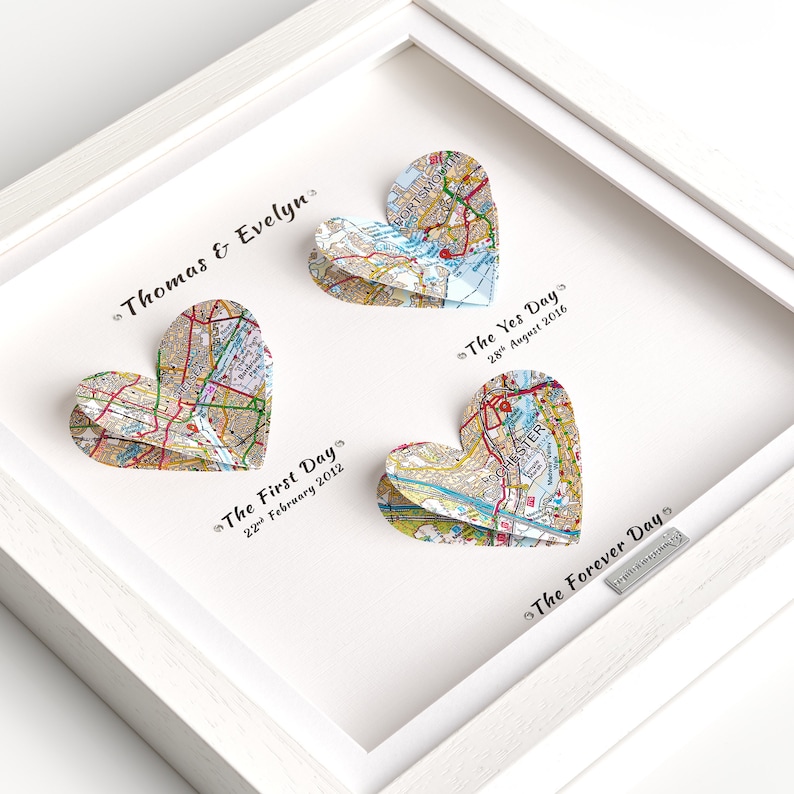 May include: A white frame with three paper map hearts. The hearts are folded to create a 3D effect. The text on the hearts reads "The First Day 22 February 2012", "The Yes Day 28 August 2016", and "The Forever Day". The names "Thomas & Evelyn" are written at the top of the frame.