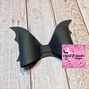 May include: Black bat wing hair bow with a black bow in the center. The bow is made of felt and has a slightly textured finish. The bow is attached to a clip.