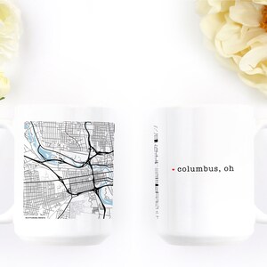 May include: Two white ceramic mugs. One mug features a black and white map of Columbus, Ohio, with a light blue river. The other mug has the text "columbus, oh" with a red dot. White flowers are in the background.