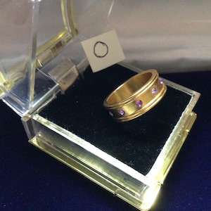 Op de afbeelding: Een goudkleurige ring met een bandontwerp, met een rij kleine, ronde, paarse edelstenen. De ring wordt gepresenteerd in een transparante, scharnierende doos met een zwart fluwelen interieur. Een klein, wit label met een getekende cirkel is ook zichtbaar.
