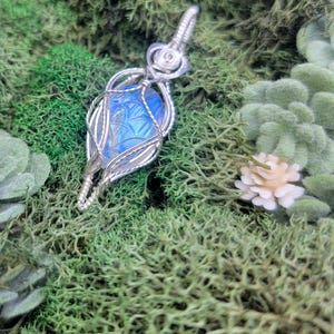 May include: A silver wire-wrapped pendant with a blue, carved gemstone. The pendant has an intricate design, with the gemstone held within a cage-like structure. The background features green moss and small white flowers, creating a natural aesthetic.