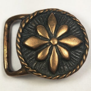 May include: A vintage bronze belt buckle with a raised, stylized flower design. The buckle has a rope-like border and a dark brown finish.