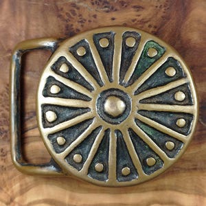 May include: A vintage brass belt buckle with a circular design. The buckle features a central circle with radiating triangular shapes, each adorned with small circular accents. The metal has a weathered appearance with hints of green patina.