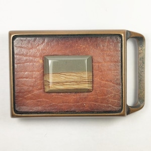 May include: A brass belt buckle with a leather inlay and a square stone with brown and gray stripes in the center.
