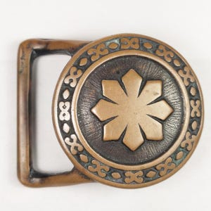 May include: A circular, bronze-colored belt buckle with an intricate design. The buckle features a central eight-pointed star surrounded by a decorative border.