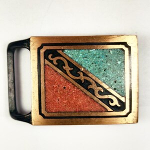 May include: A square belt buckle with a black metal frame and a copper colored background. The buckle is decorated with a geometric design of turquoise and red stones.
