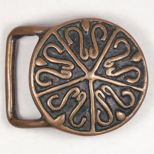 May include: A bronze belt buckle with a circular design featuring a repeating pattern of stylized S shapes. The buckle is a dark bronze color with a slightly aged patina.