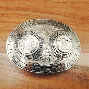May include: Silver-toned oval belt buckle with intricate detailing. Features two circular designs with a Native American profile and a bison. The buckle has a decorative border and floral accents, suitable for Western-style attire.