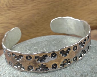 May include: A silver cuff bracelet with a detailed pattern of geometric shapes and circles. The bracelet has a slightly tarnished, aged appearance, and is displayed on a gray stone. The design features a series of repeating motifs.