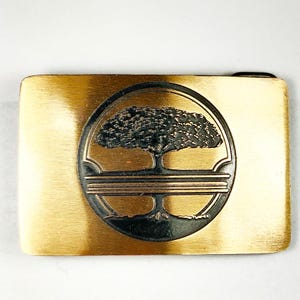 May include: A gold-toned metal belt buckle with a black and gold tree design. The tree is surrounded by a circle with three concentric rings.