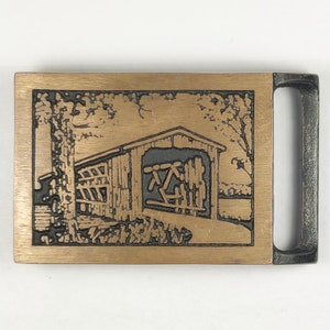 May include: A brass belt buckle with a detailed image of a covered bridge. The buckle is rectangular with a smooth, brushed finish. The bridge is depicted in a rustic style with wooden beams and a gabled roof.