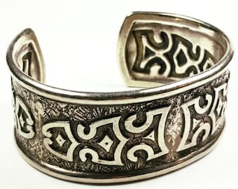 May include: A silver cuff bracelet with an intricate geometric design. The bracelet is made of a smooth, polished metal and has a slightly textured surface.