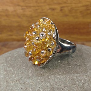 May include: A silver ring with a large, oval-shaped cluster of yellow and clear beads. Small silver beads are interspersed throughout the cluster. The ring is set on a gray stone surface.