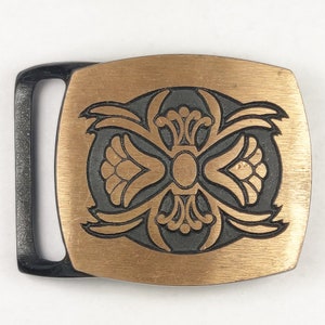 May include: A gold-toned belt buckle with a black, intricate design in the center. The buckle is rectangular and has a brushed finish.