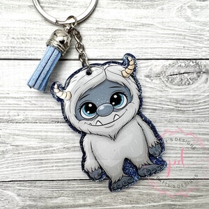 May include: A blue and white glitter keychain featuring a cartoon Yeti with big eyes and a toothy grin. The keychain has a blue tassel.