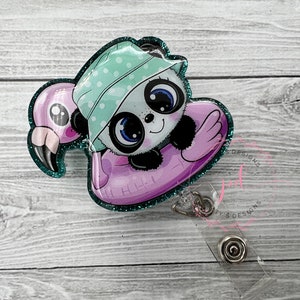 May include: A teal glitter badge reel with a panda wearing a green and white polka dot hat and riding a pink flamingo float. The reel has a clear plastic clip and a silver swivel.