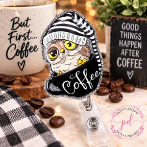May include: A badge reel featuring a cartoon owl with large yellow eyes wearing a black and white striped nightcap, sitting inside a black coffee mug with the word Coffee written in white script, all outlined with a silver glitter border.