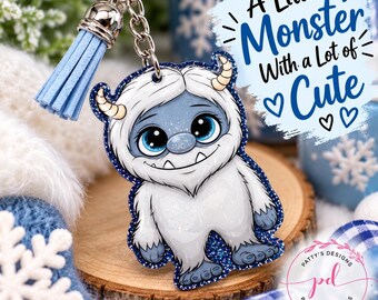 May include: A keychain featuring a white, furry monster character with blue eyes, small horns, and a glittery dark blue border. It includes a light blue tassel and a silver metal ring. The text reads, A Little Monster With a Lot of Cute.