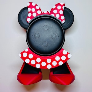 Może przedstawiać: Czarny głośnik inteligentny z akcesorium w stylu Minnie Mouse. Akcesorium zawiera czerwoną kokardę w białe grochy, czarne uszy, czerwony kołnierz w białe grochy i czerwone buty. Głośnik posiada przyciski sterowania.