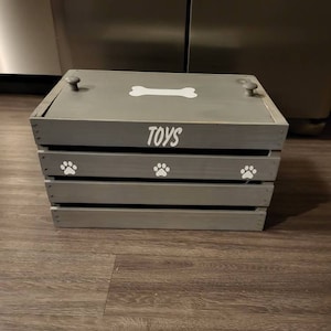 Puede incluir: Una caja de juguetes de madera gris con un dise&ntilde;o de hueso blanco y huellas de patas. La caja tiene una tapa y tres compartimentos. La tapa tiene dos pomos grises. La caja est&aacute; etiquetada como "TOYS".